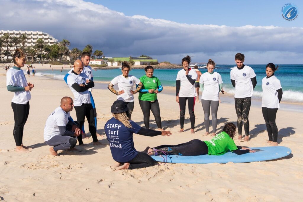 Fuertetribu Clase Surf Principiante 1 1024x683 El monitor de surf explica la posición correcta en la tabla en freinte de sus estudiantes en la playa.
