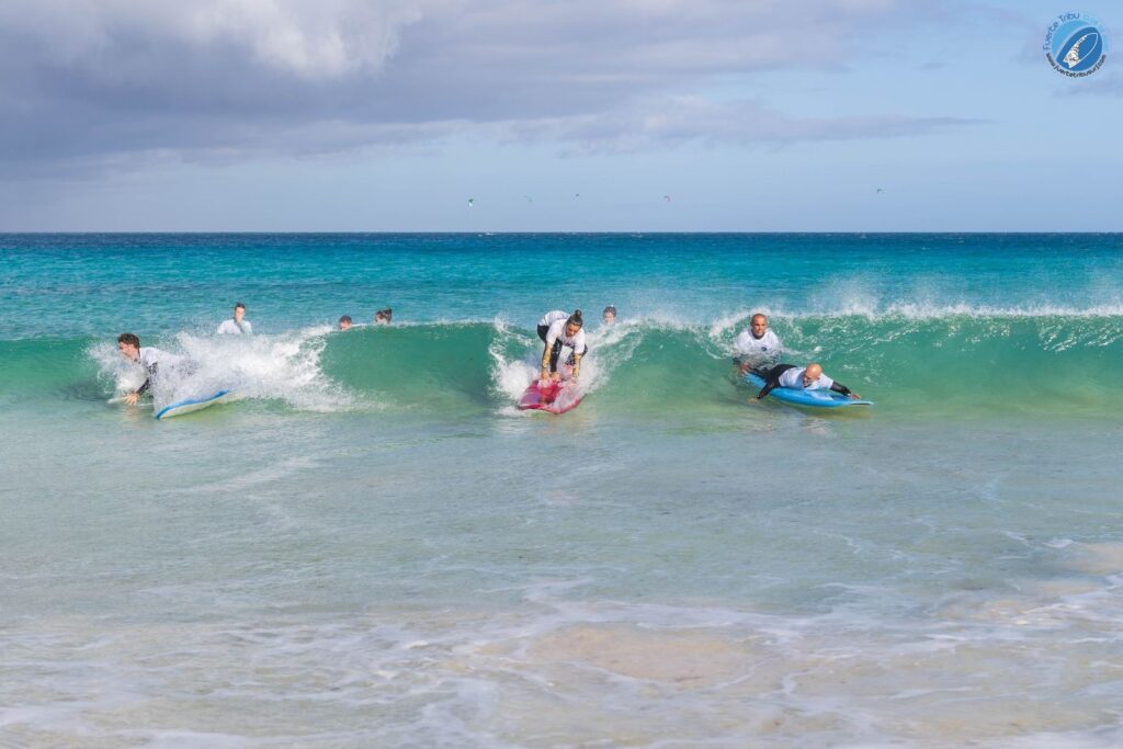 Fuertetribu Clase Surf Principiante 3 1024x683 Varias personas estan cogiendo una ola intentando a levantarse en la tabla.