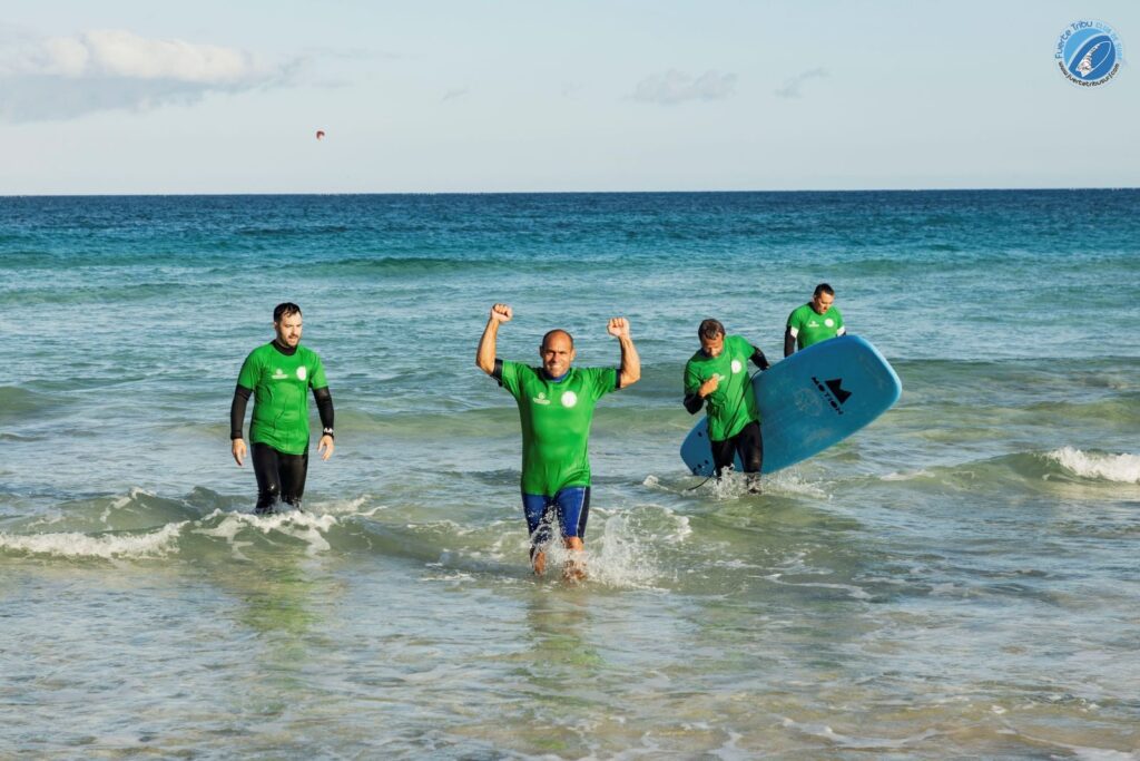 Fuertetribu Clase Surf Principiante 5 1024x683 Un grupo de principiantes sale del agua con una tabla del surf lleno de alegría.