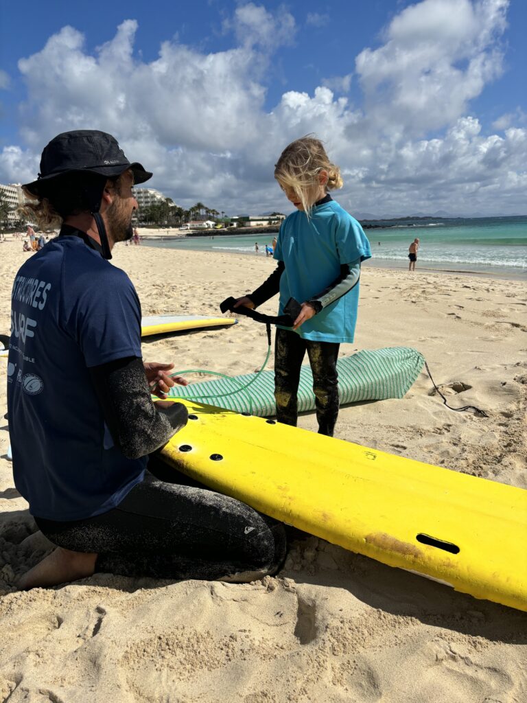 Fuertetribu Surf Para Ninos 1 768x1024 Niña y su monitor de surf preparando su tabla de surf en una playa en el norte de Fuerteventura.