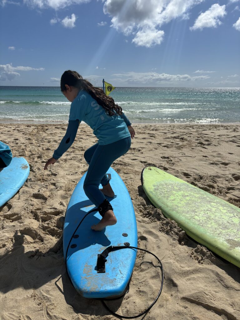 Fuertetribu Surf Para Ninos 7 768x1024 Niña practicando su posición del surf con una tabla en la arena en una playa cerca de Corralejo.