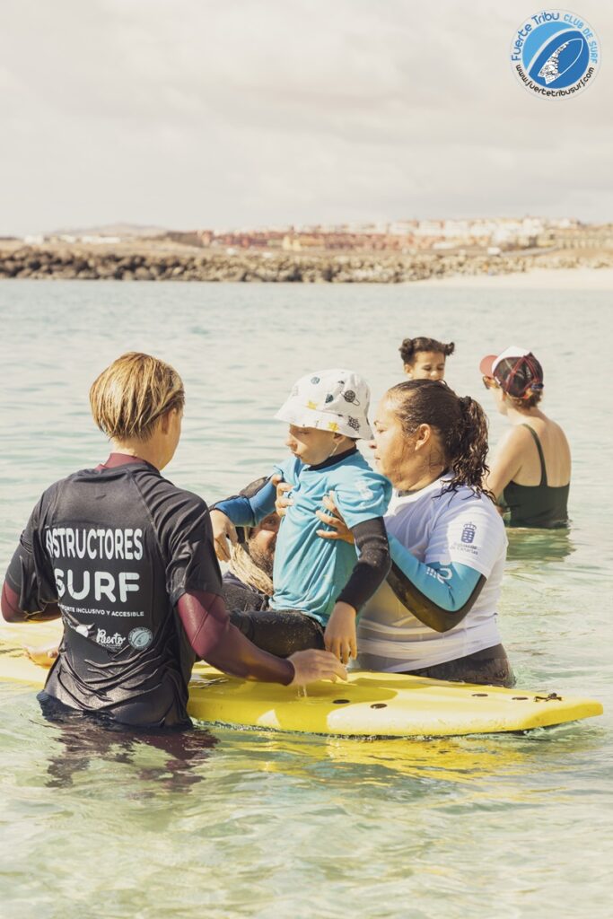 Fuertetribu Voluntariado 5 683x1024 Un voluntario ayuda a un monitor de surf a sentar por primera vez a un niño con diversidad funcional en la tabla.