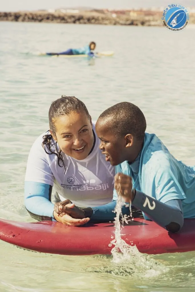 Fuertetribu Voluntariado 4 683x1024 Una voluntaria ayuda a un niño a tumbarse sobre la tabla de surf y ambos ríen felices.