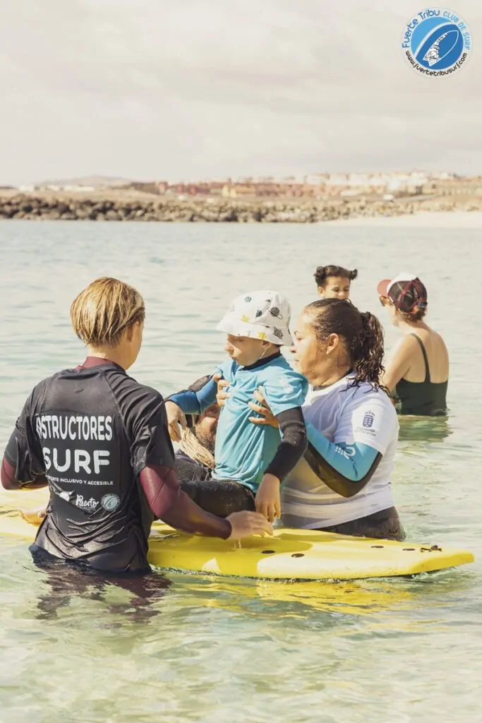 Fuertetribu Voluntariado 5 683x1024 Un voluntario ayuda a un monitor de surf a sentar por primera vez a un niño con diversidad funcional en la tabla.
